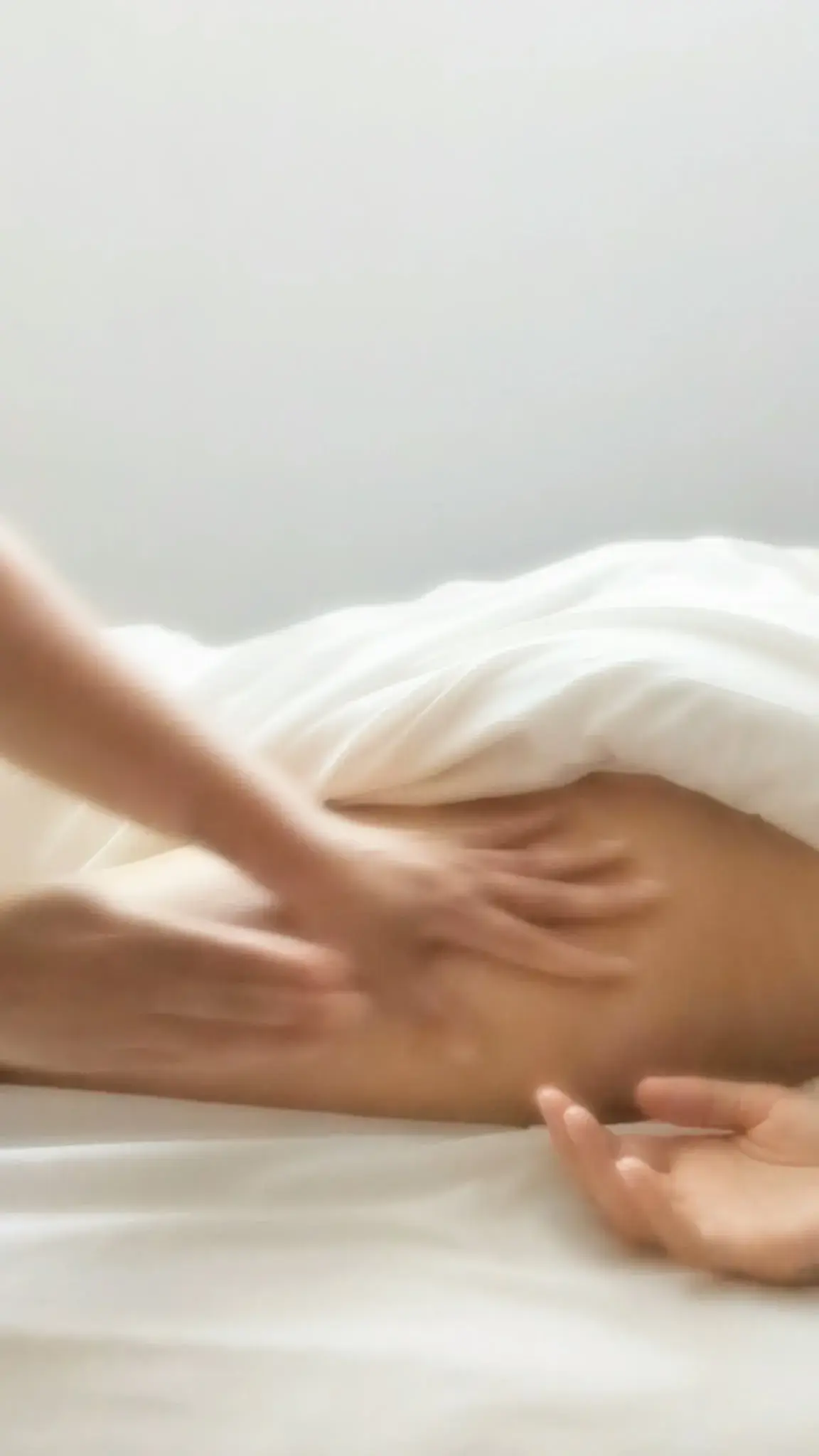 A therapist's hands working along a client's thigh beneath a white sheet, in soft focus