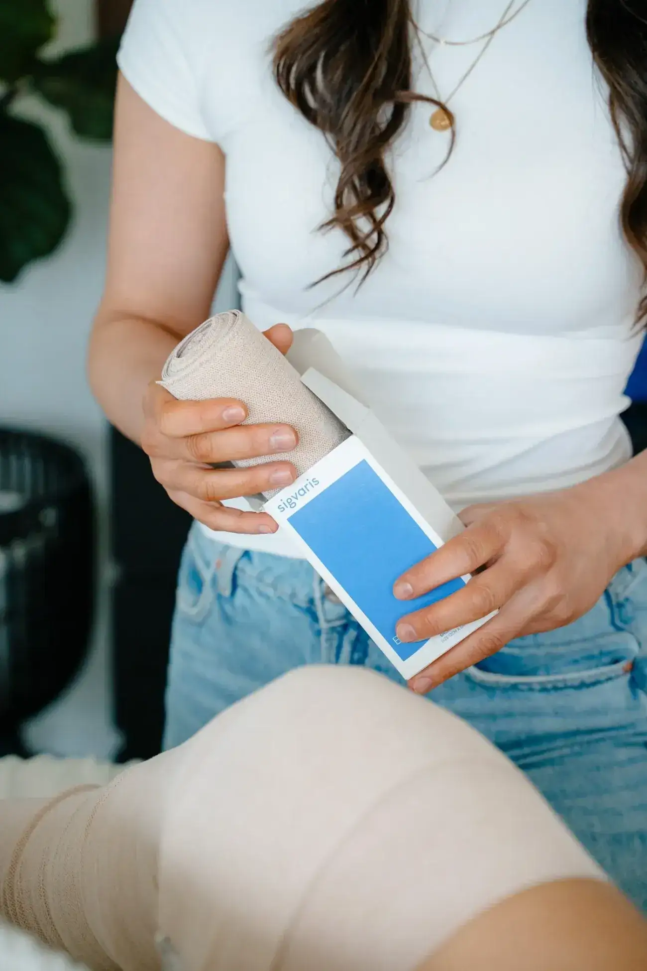 A Renu therapist holds a roll of short-stretch compression bandage and its box beside a client's bandaged leg during a compression fitting