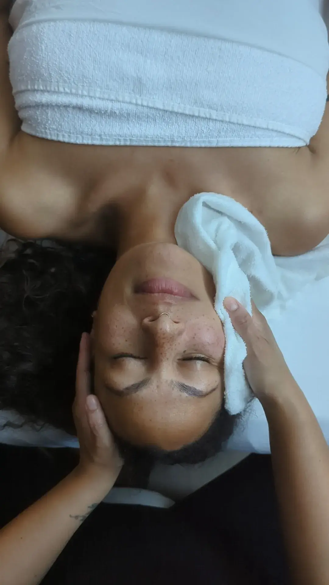 A client lying back with eyes closed while a therapist holds a folded white towel against the side of her face and jaw