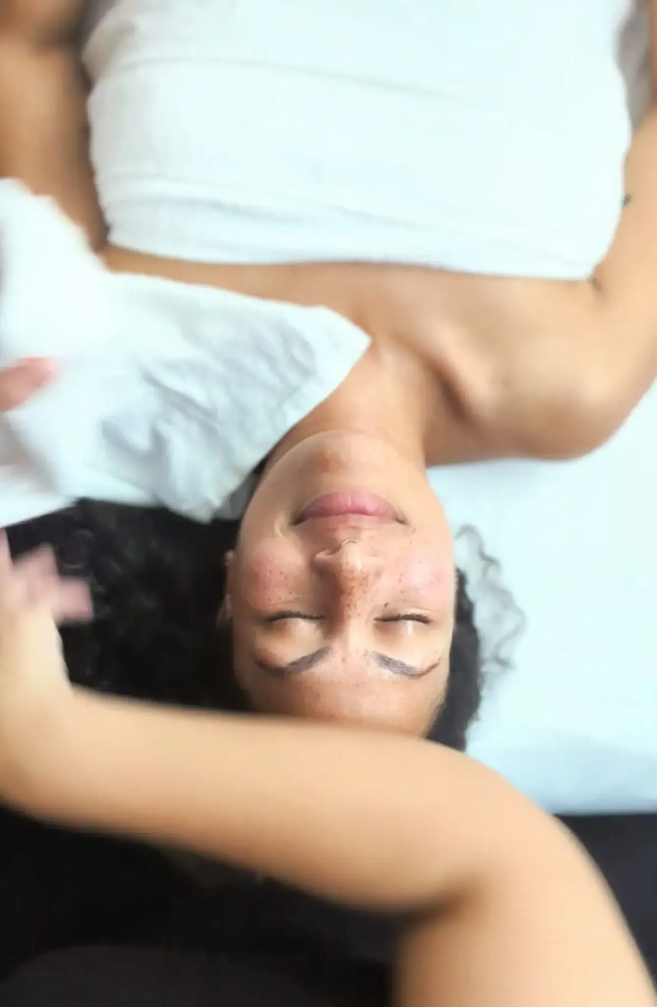 A client wrapped in a white towel lies face up, eyes closed, as a soft cloth is held beside her face during an express facial