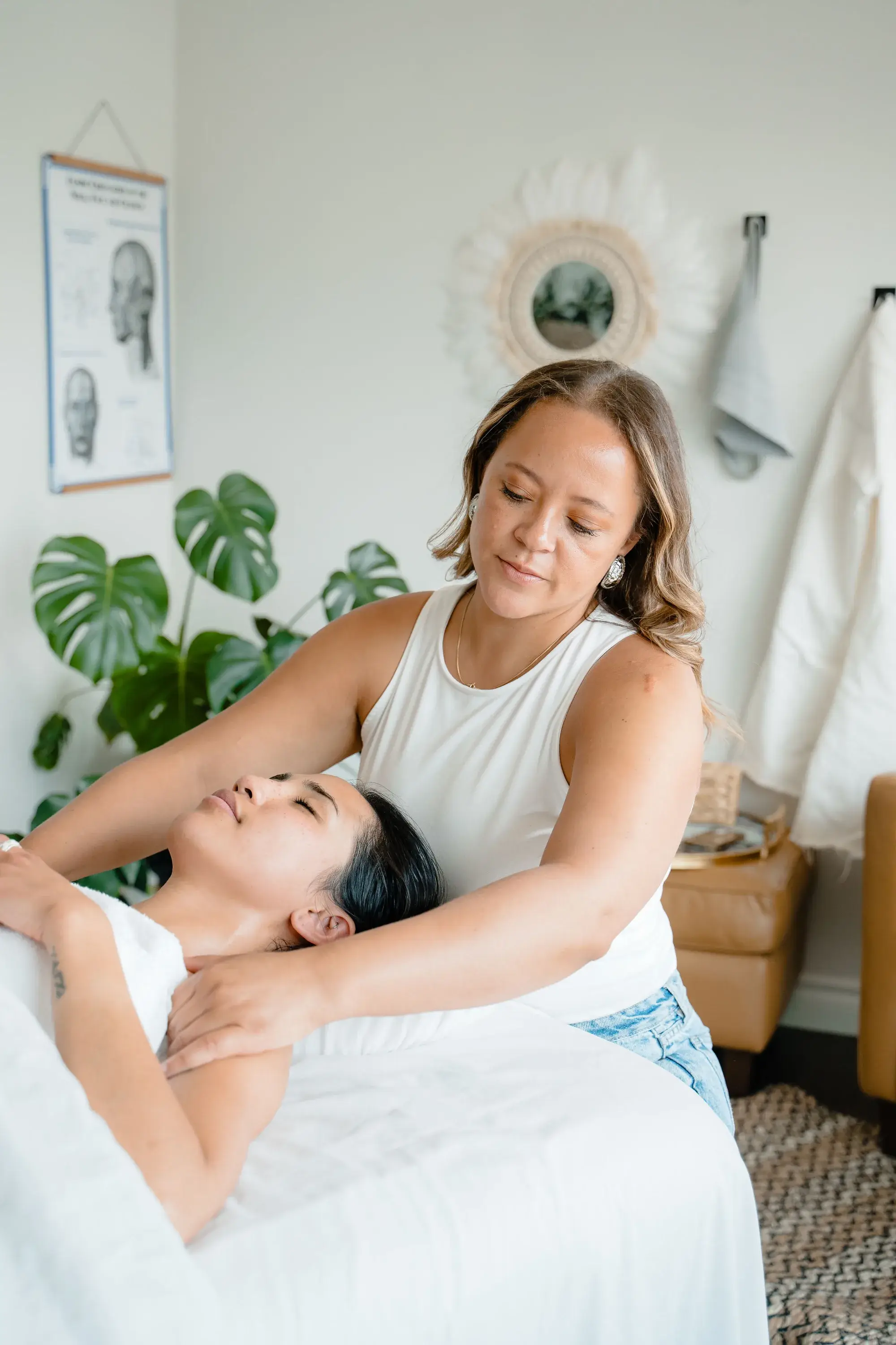 A Renu therapist resting flat hands on the neck and shoulder of a client lying face up on the treatment table, with anatomy charts and a monstera behind her