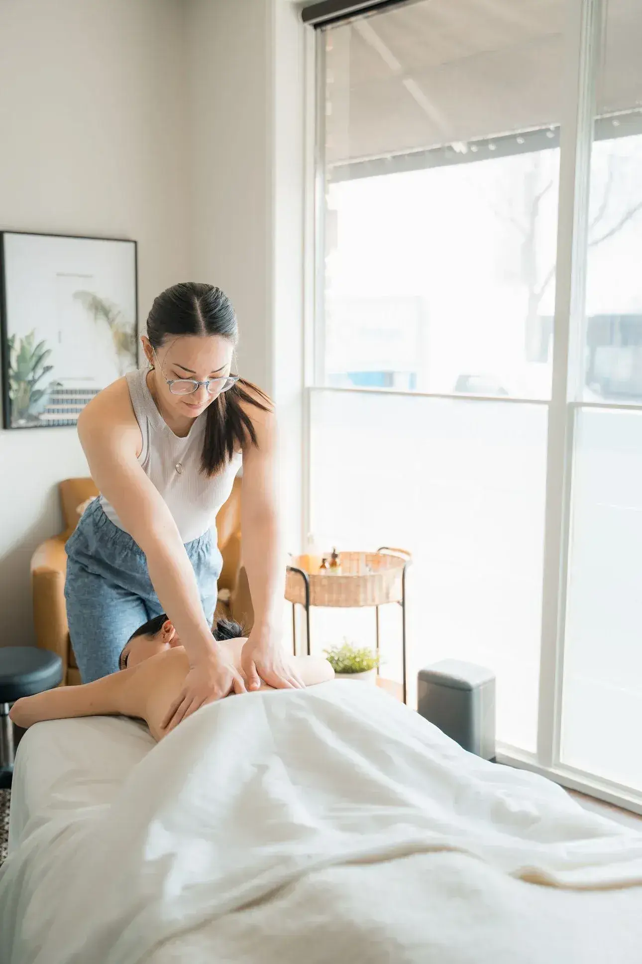 A Registered Massage Therapist working slowly across the shoulders of a client lying face down beside a bright window at Renu