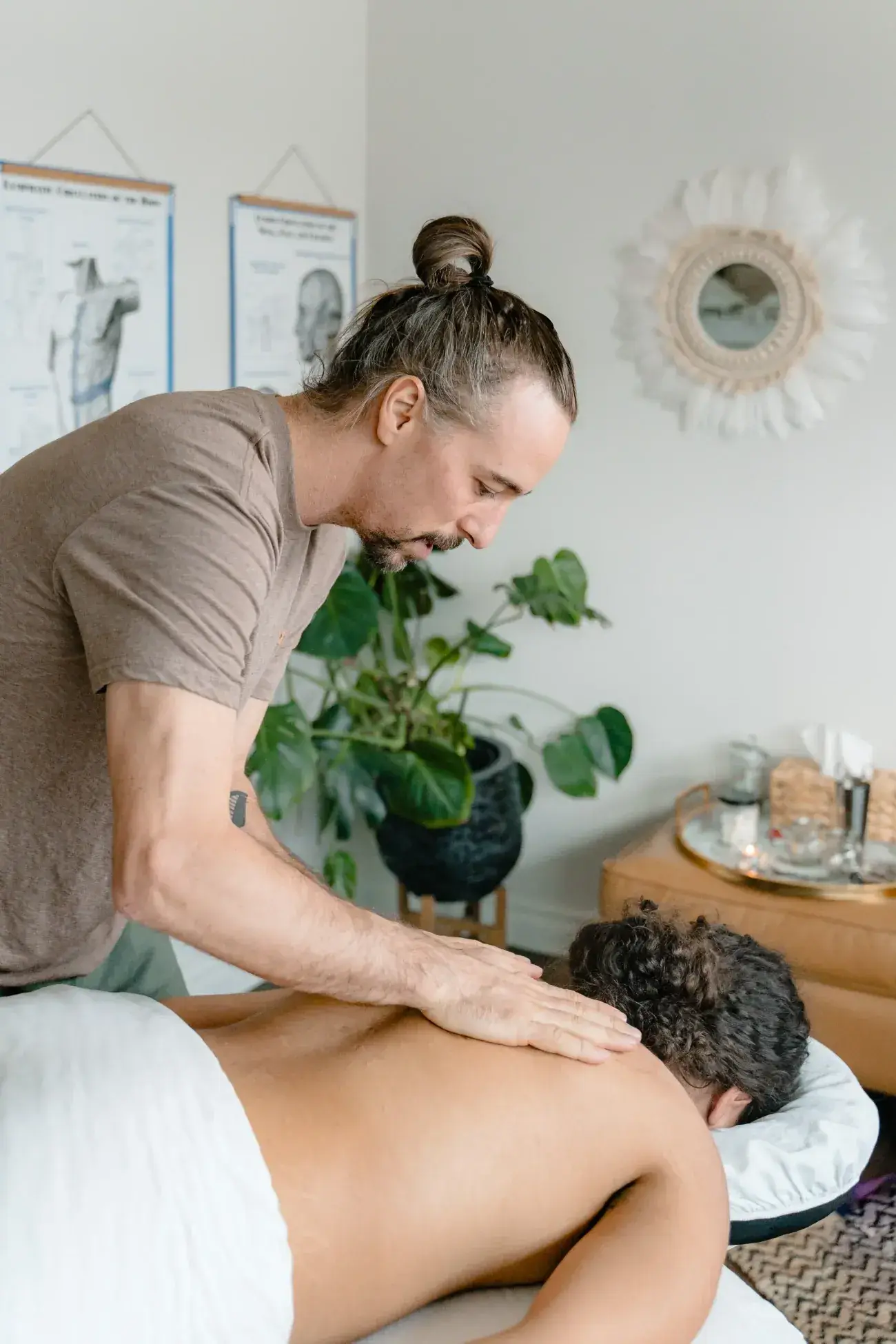 A Renu Registered Massage Therapist sweeping a flat hand along the upper back of a client lying face down, with anatomy charts on the wall behind him