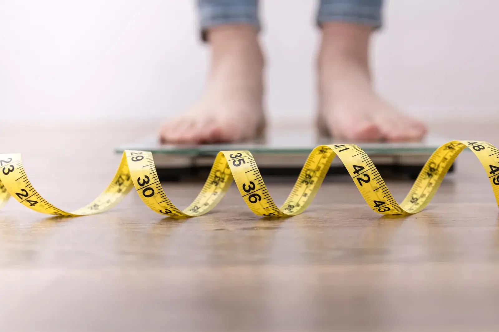 Bare feet on a bathroom scale behind a yellow tape measure curled across the floor
