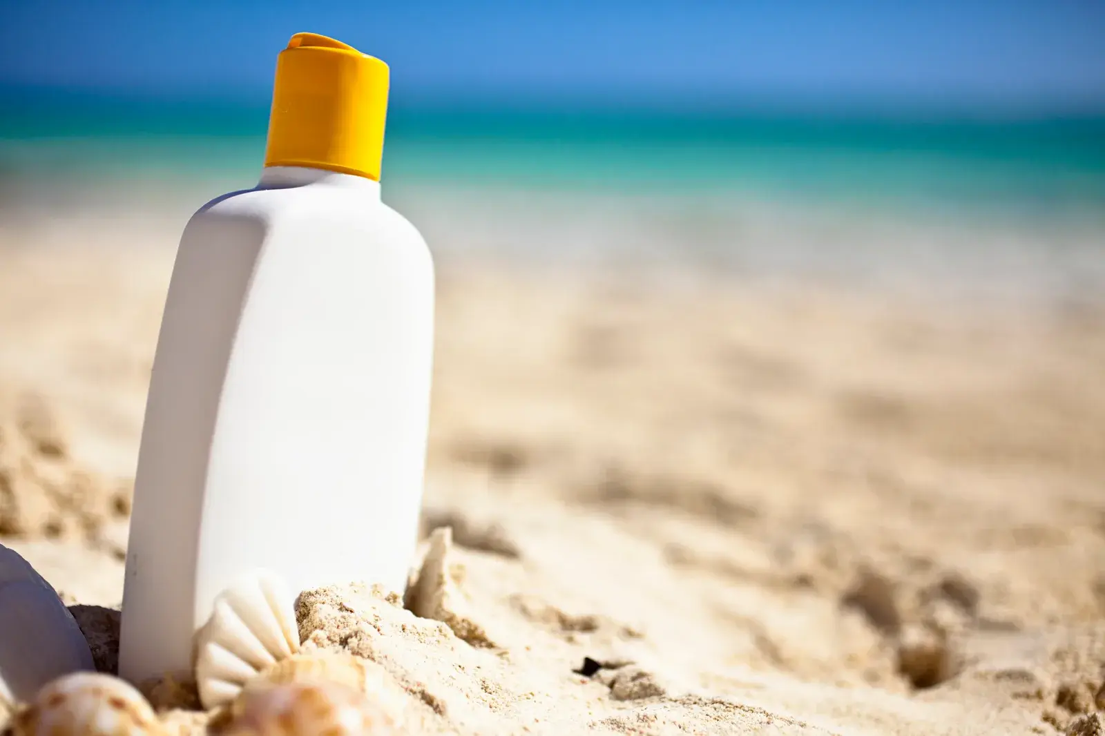 A white sunscreen bottle with an orange cap standing in beach sand with the sea behind