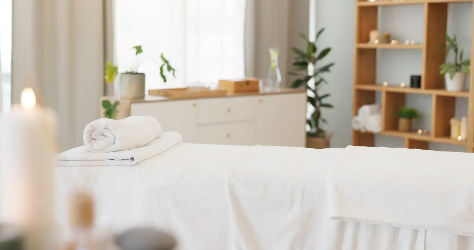A calm massage room with a made-up treatment table, a rolled towel and a lit candle in soft daylight