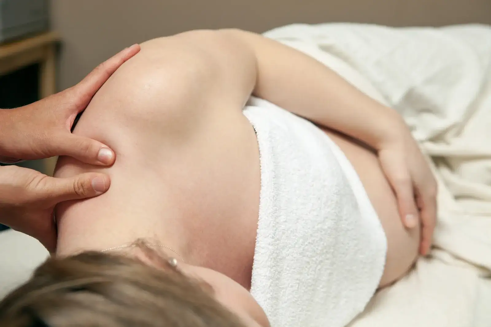 A pregnant woman lying on her side under a white towel while a therapist presses into the muscle at the top of her shoulder