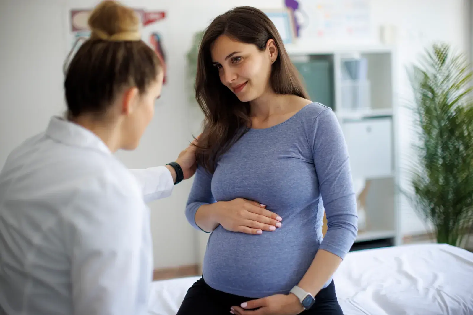 A pregnant woman in a grey top sits on an examination couch with one hand on her belly while a clinician in a white coat talks with her