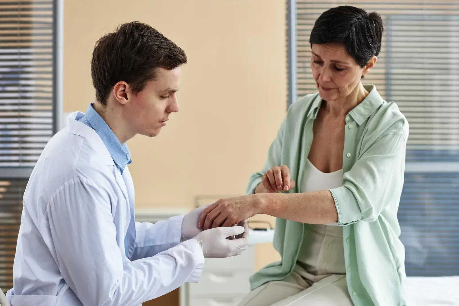 A doctor in gloves examines the skin on a woman's forearm during a clinic visit, checking for signs of psoriasis