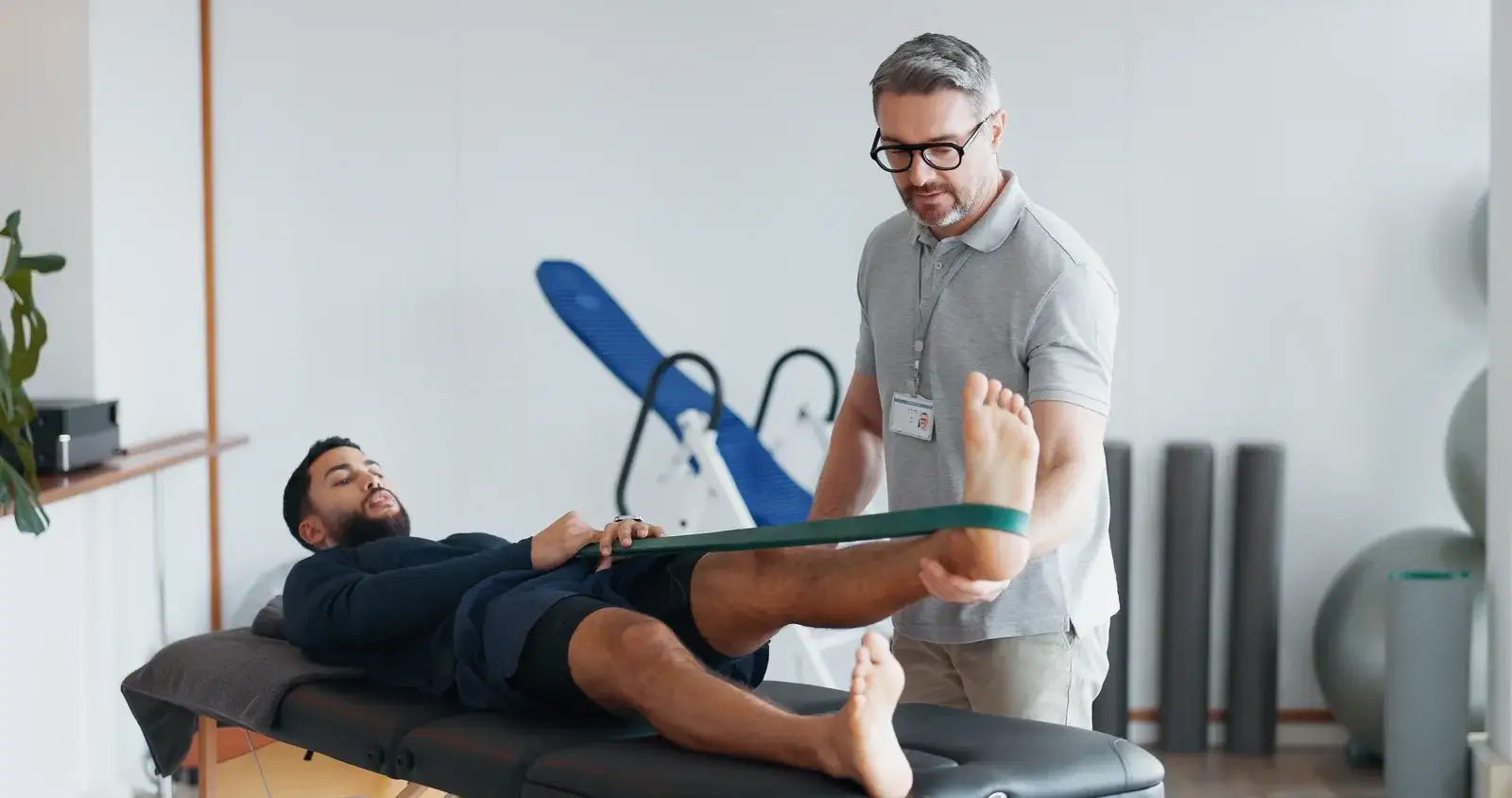 A clinician guiding a man lying on a treatment table through a leg exercise with a green resistance band