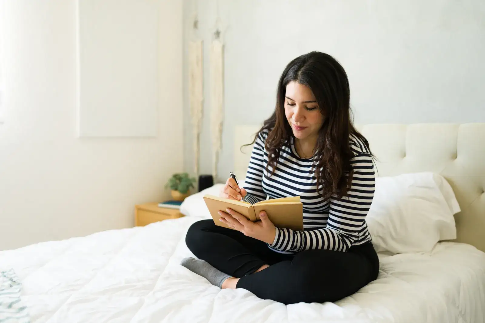 A woman sitting cross-legged on a white bed, writing in a notebook in soft daylight