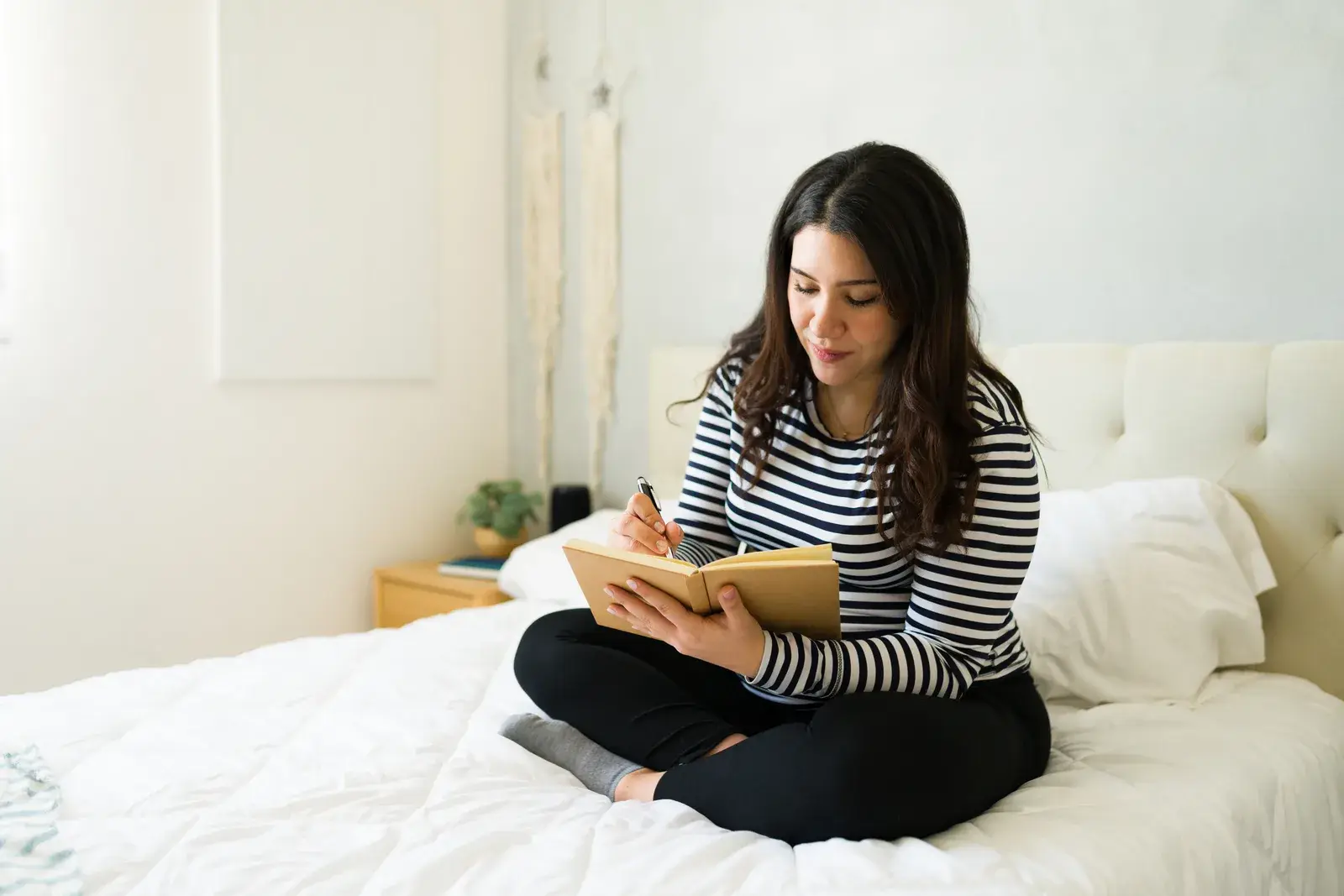 A woman sitting cross-legged on a white bed, writing in a notebook in soft daylight