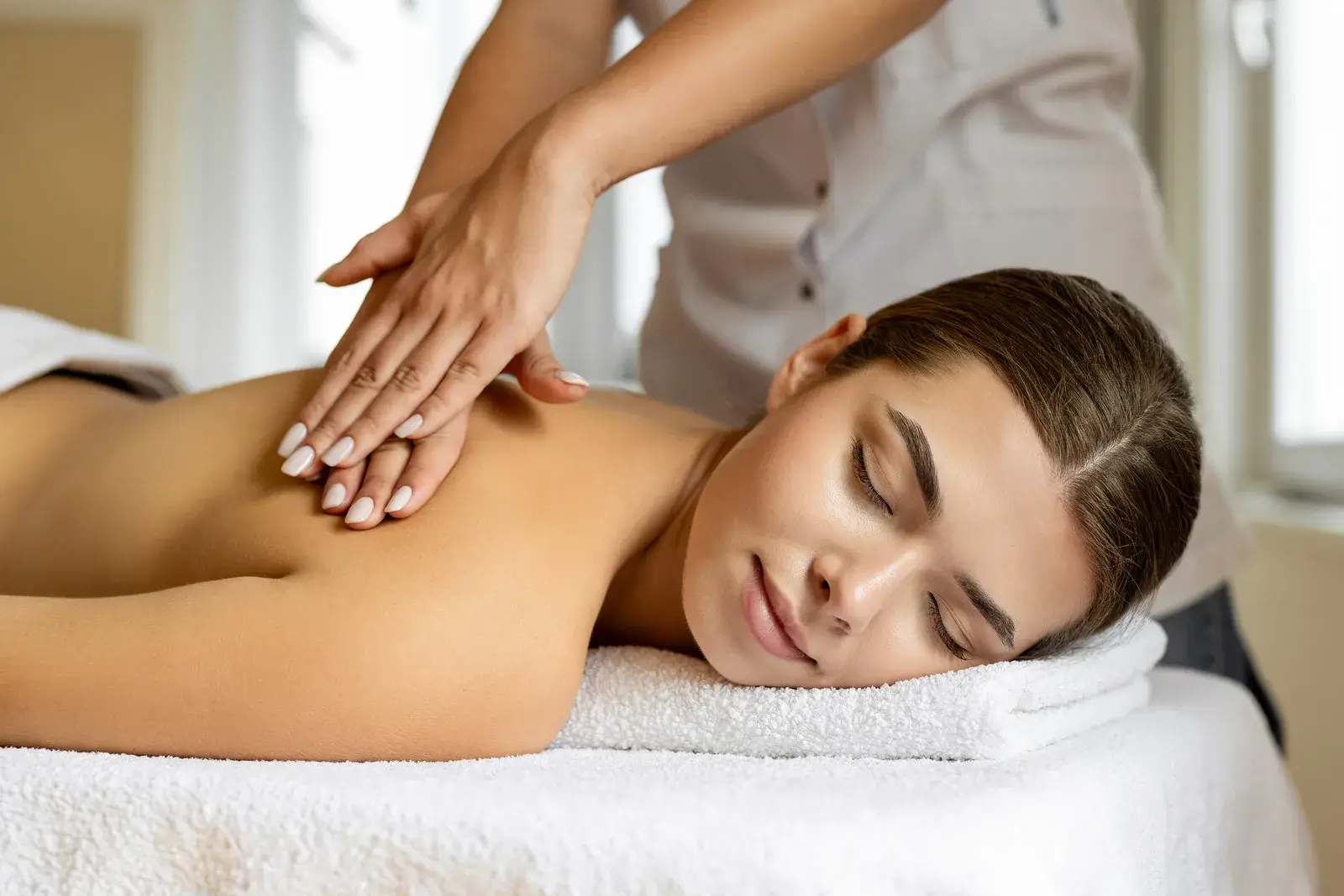 A woman resting with her eyes closed on a towel-covered table while a therapist's hands stroke her back