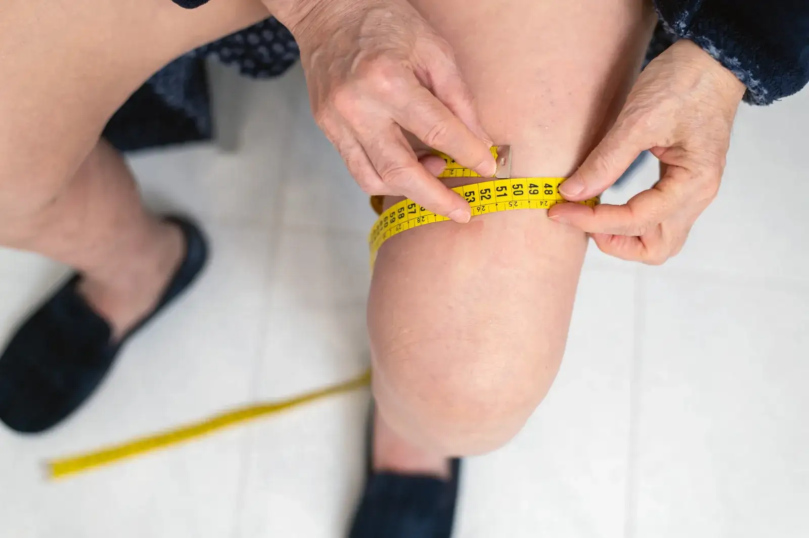 Hands wrapping a yellow tape measure around a bare leg just above the knee to measure its girth