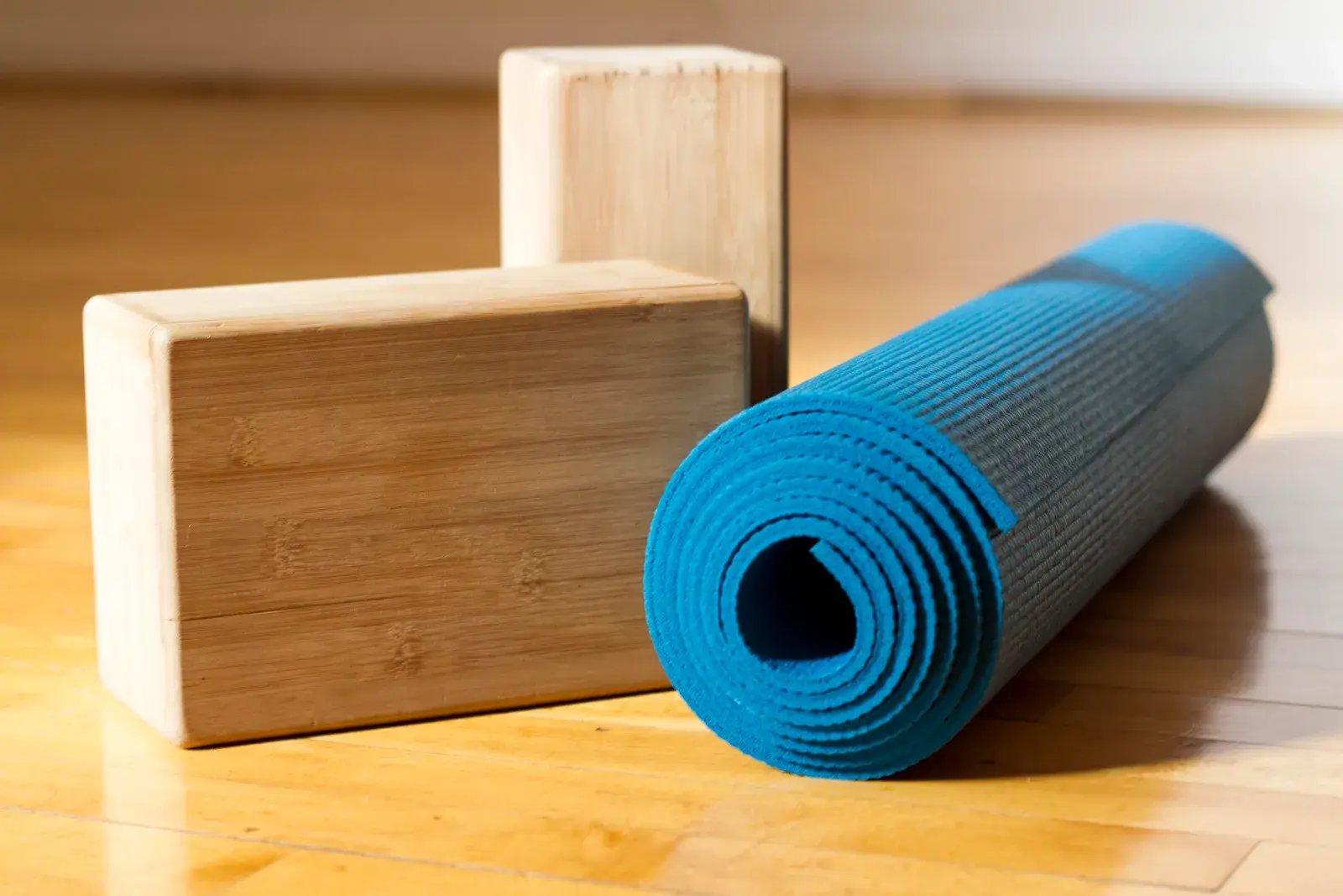A rolled blue yoga mat and two wooden yoga blocks on a wooden studio floor