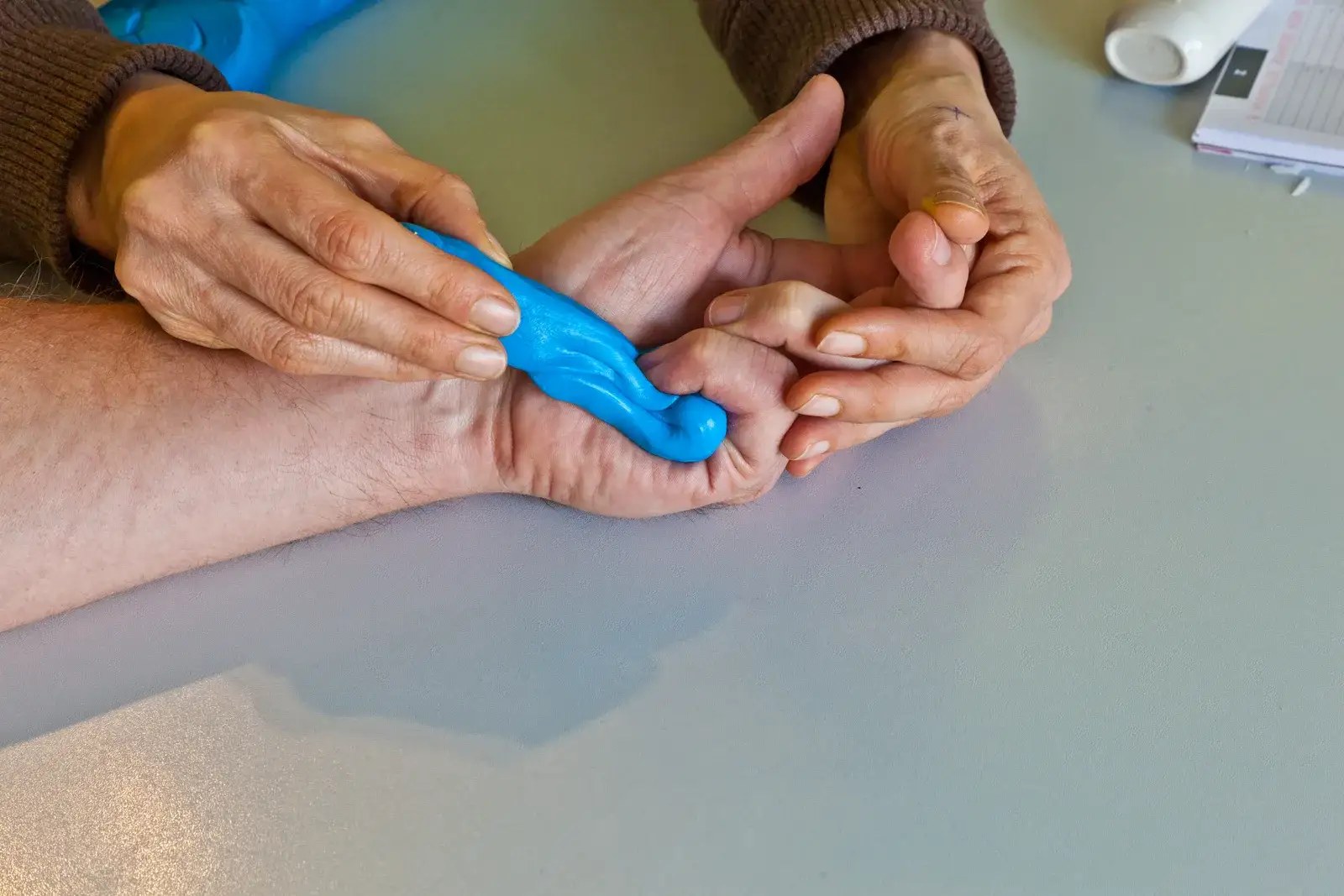 A therapist guiding an older person's fingers around a blue hand-therapy tool to work stiff, arthritic joints