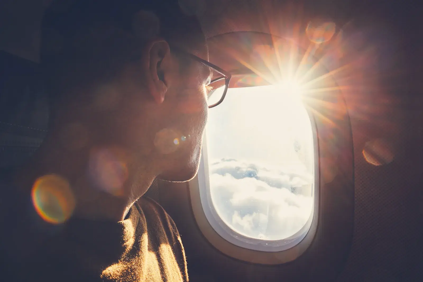 A passenger in glasses looking out of an airplane window at sunlight over the clouds