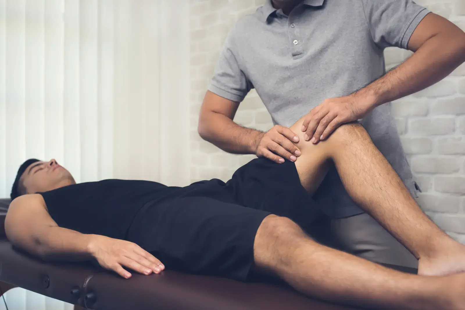 A therapist's hands working on the front of the thigh above the knee of a man lying face up on a treatment table