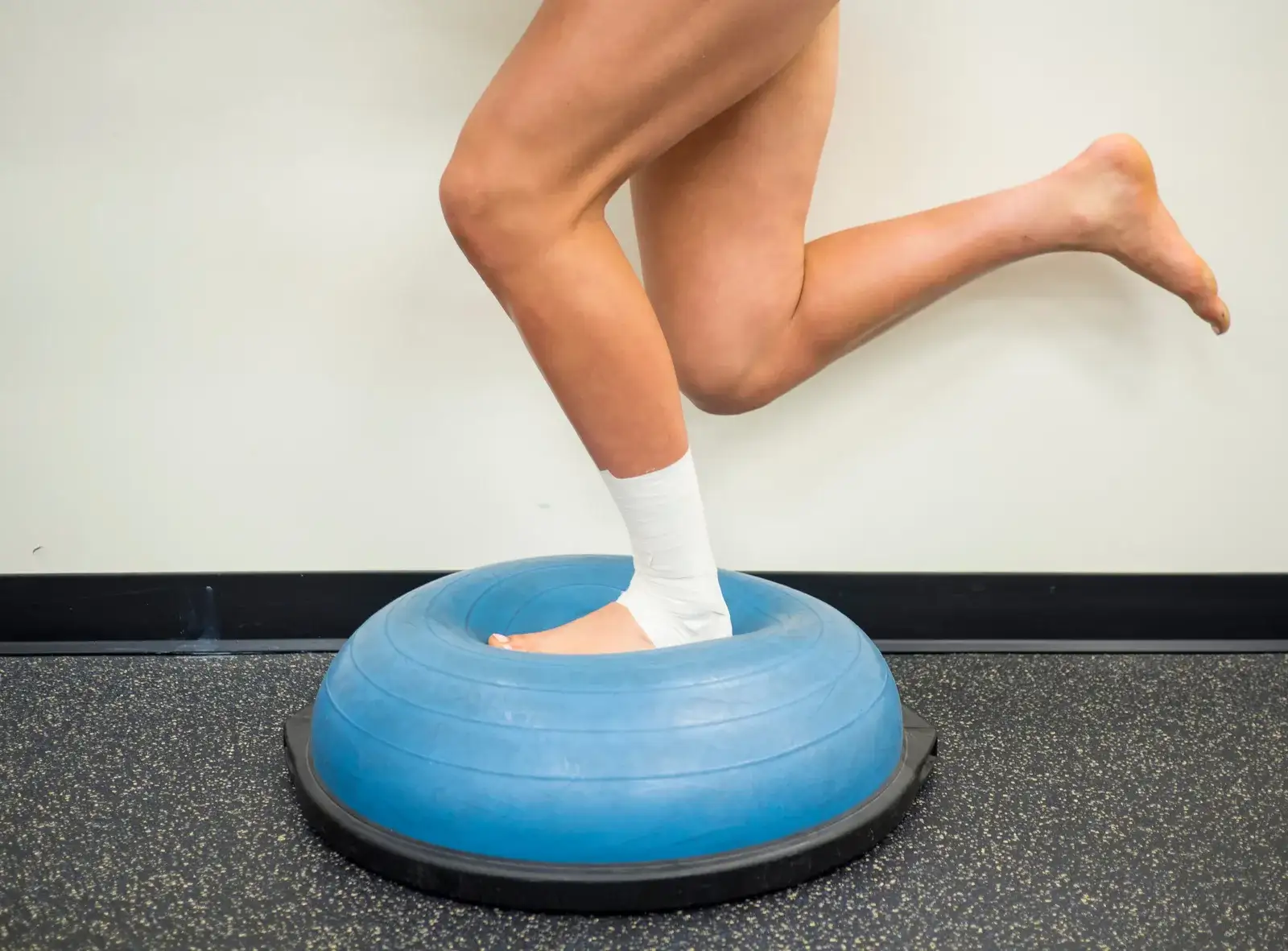 An athlete balancing on one bare foot on a half-ball balance trainer in a gym during ankle rehabilitation