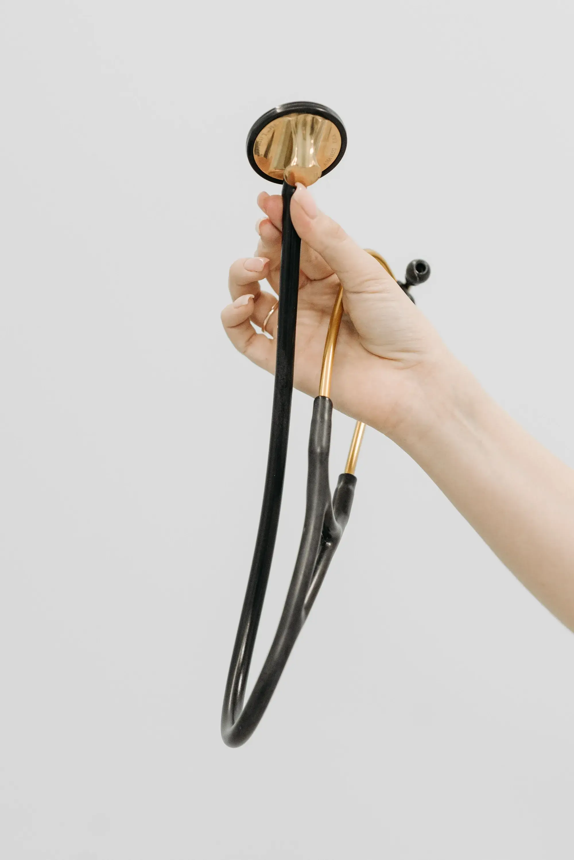 A hand holding up a black and gold stethoscope against a plain white wall