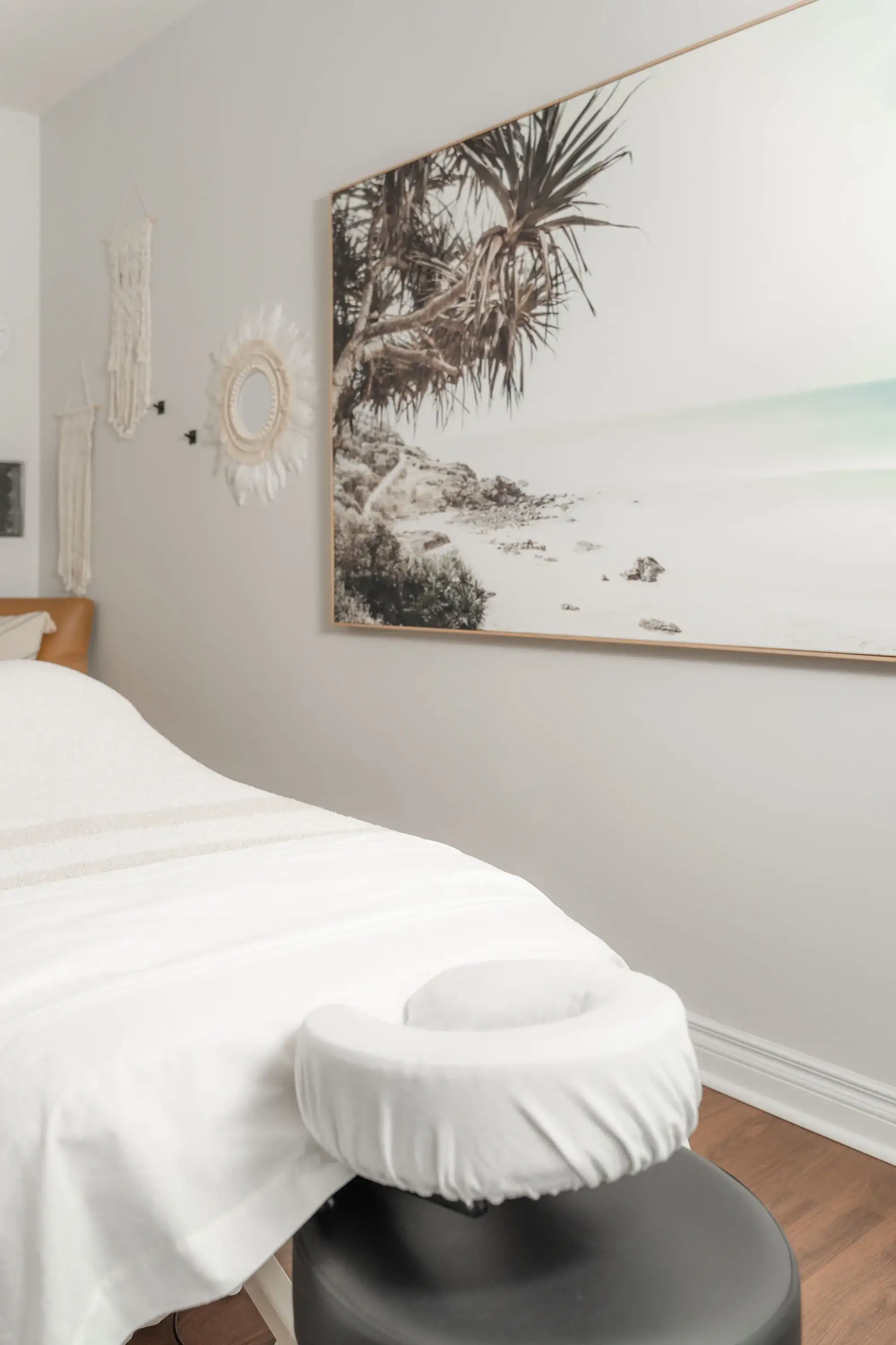 A treatment table dressed in white linen with a padded face cradle, beneath a large framed photograph of a pandanus tree on a beach, at Renu